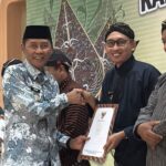 Men in traditional batik and kebaya-like attire shake hands on stage while receiving certificates at an awards ceremony.