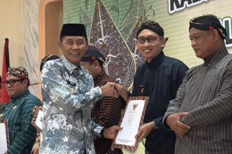 Men in traditional batik and kebaya-like attire shake hands on stage while receiving certificates at an awards ceremony.