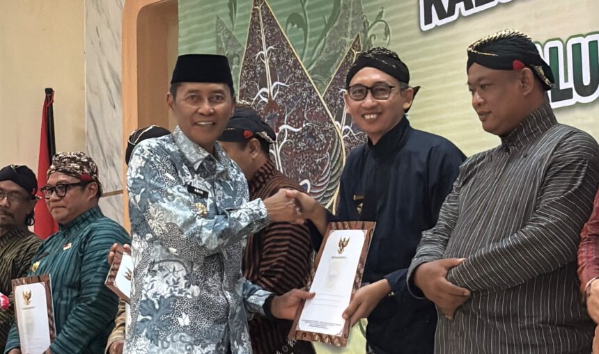 Men in traditional batik and kebaya-like attire shake hands on stage while receiving certificates at an awards ceremony.