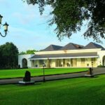 Park scene featuring a white pavilion-like building with arches, black lamp posts, and a green lawn on a sunny day