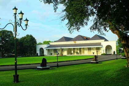 Park scene featuring a white pavilion-like building with arches, black lamp posts, and a green lawn on a sunny day
