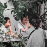 Three people sit at a shaded outdoor table under large tropical leaves, chatting with drinks on the table, seen from behind one person.