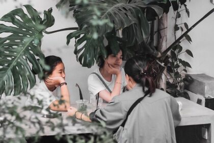 Three people sit at a shaded outdoor table under large tropical leaves, chatting with drinks on the table, seen from behind one person.