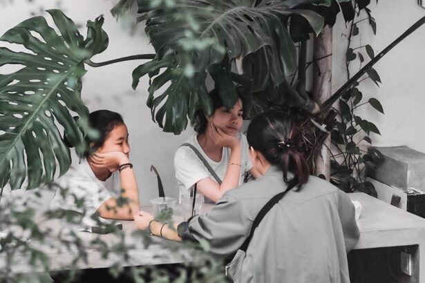Three people sit at a shaded outdoor table under large tropical leaves, chatting with drinks on the table, seen from behind one person.