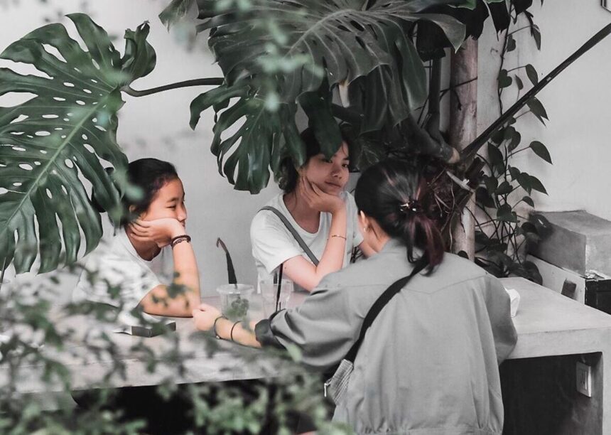 Three people sit at a shaded outdoor table under large tropical leaves, chatting with drinks on the table, seen from behind one person.