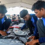 Four boys in blue uniforms sit on a black plastic sheet under a tent, focused on a hands-on project.