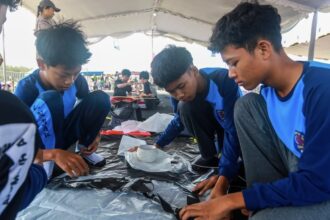 Four boys in blue uniforms sit on a black plastic sheet under a tent, focused on a hands-on project.