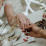 Couple exchanging rings at a wedding table, hands joined near a bouquet and glassware on a decorated table.