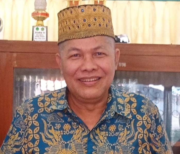 Smiling man wearing a gold ceremonial hat and blue batik shirt indoors.
