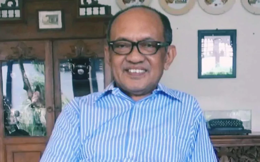 Smiling man with glasses in a blue striped shirt seated indoors, with a wooden cabinet and framed photos behind him.
