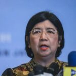 Woman with short black hair and glasses speaks at a press conference, microphones in front of her against a blue backdrop.