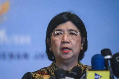 Woman with short black hair and glasses speaks at a press conference, microphones in front of her against a blue backdrop.