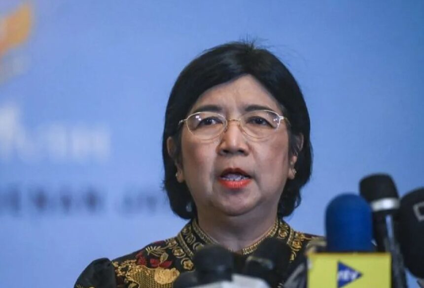 Woman with short black hair and glasses speaks at a press conference, microphones in front of her against a blue backdrop.