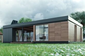 Modern single-story house with wooden cladding and large glass doors, set on a green lawn with a cloudy sky above.