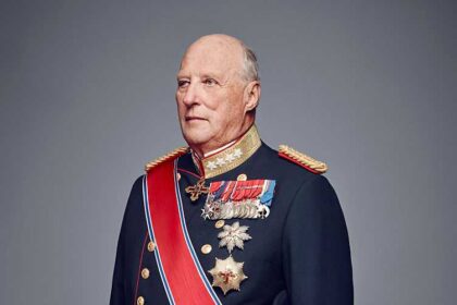 Older man in ceremonial military dress uniform with medals, sash, and sword, standing against a grey backdrop.