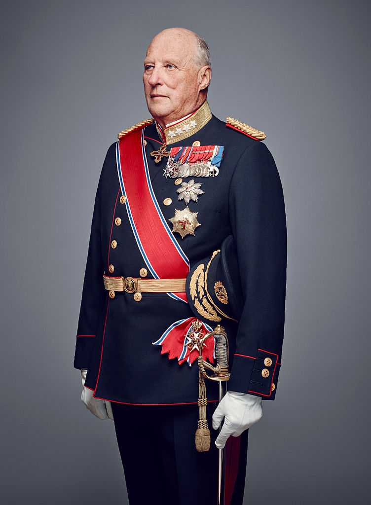 Older man in ceremonial military dress uniform with medals, sash, and sword, standing against a grey backdrop.