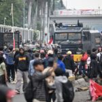 Crowd of protesters in a street, facing a black armored police vehicle with blue lights; red flags and barriers nearby.