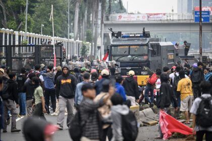 Crowd of protesters in a street, facing a black armored police vehicle with blue lights; red flags and barriers nearby.