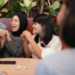 Four women in hijabs sit at a cafe table, smiling as one holds cash and the others cheer or clap, suggesting a money-related exchange or celebration.