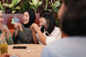 Four women in hijabs sit at a cafe table, smiling as one holds cash and the others cheer or clap, suggesting a money-related exchange or celebration.