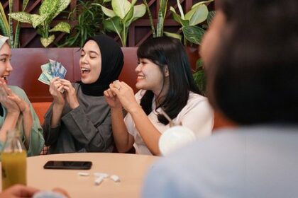 Four women in hijabs sit at a cafe table, smiling as one holds cash and the others cheer or clap, suggesting a money-related exchange or celebration.