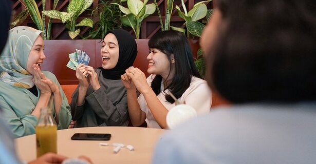 Four women in hijabs sit at a cafe table, smiling as one holds cash and the others cheer or clap, suggesting a money-related exchange or celebration.