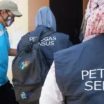 Census officers in dark vests marked PETUGAS SENSUS approach a doorway while a man in a blue shirt wearing a face mask watches nearby.