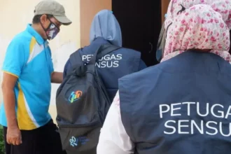 Census officers in dark vests marked PETUGAS SENSUS approach a doorway while a man in a blue shirt wearing a face mask watches nearby.
