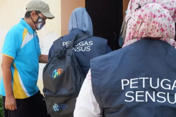 Census officers in dark vests marked PETUGAS SENSUS approach a doorway while a man in a blue shirt wearing a face mask watches nearby.