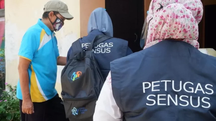 Census officers in dark vests marked PETUGAS SENSUS approach a doorway while a man in a blue shirt wearing a face mask watches nearby.