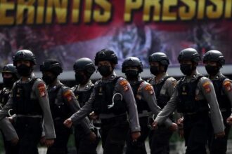 Line of armed police in helmets marching in formation during a parade, with a bold banner in the background.