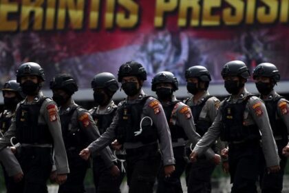 Line of armed police in helmets marching in formation during a parade, with a bold banner in the background.