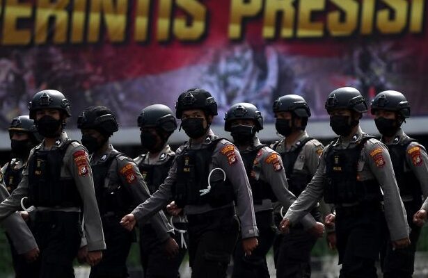 Line of armed police in helmets marching in formation during a parade, with a bold banner in the background.