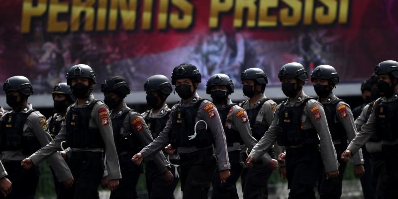 Line of armed police in helmets marching in formation during a parade, with a bold banner in the background.