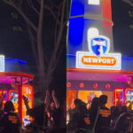 Nighttime scene at a Newport amusement booth with blue and red neon lights and a cheering crowd gathered in front.