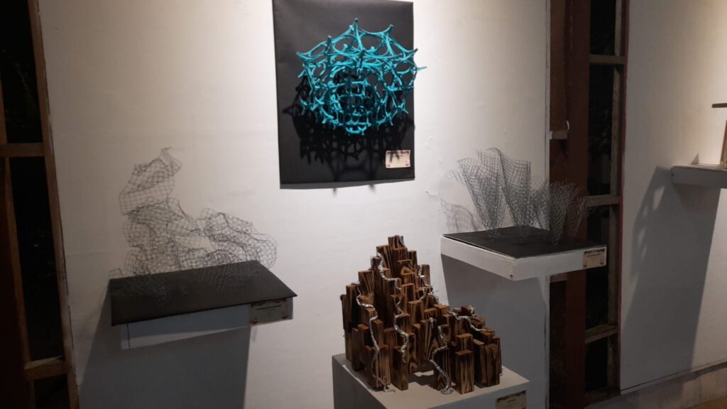Abstract turquoise knot sculpture mounted on a black panel, with wire-mesh forms on pedestals and a wooden block sculpture in the foreground in a white gallery.
