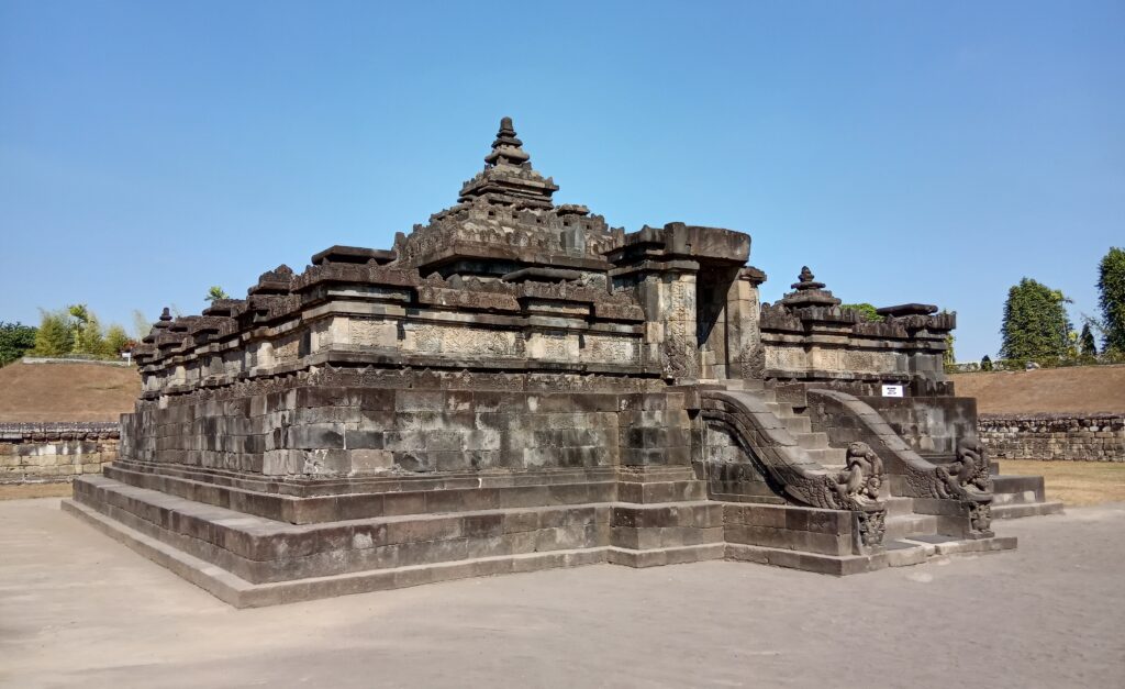 9d1b4a125ead3f12ecdc0b1dc86e4d81 - Mabur.co Stone temple with multiple tiers and a central stepped tower, decorated with carved reliefs under a clear blue sky on a raised platform.