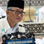 Indonesian official wearing a black peci talks into a mic at a conference, with a 'Menteri Pekerjaan Umum' nameplate in front and papers on the table.
