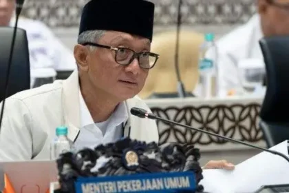 Indonesian official wearing a black peci talks into a mic at a conference, with a 'Menteri Pekerjaan Umum' nameplate in front and papers on the table.
