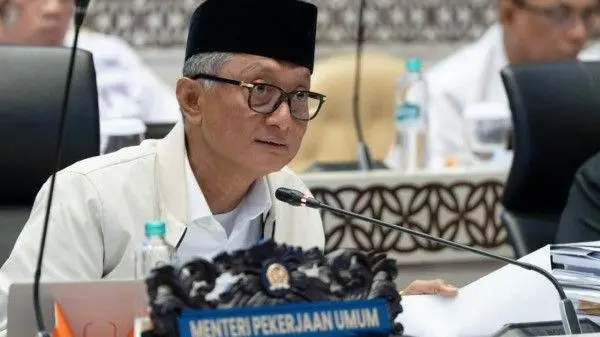 Indonesian official wearing a black peci talks into a mic at a conference, with a 'Menteri Pekerjaan Umum' nameplate in front and papers on the table.