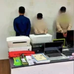 Three people with their backs to the camera stand behind a table with printers, a laptop, and office supplies in a room.