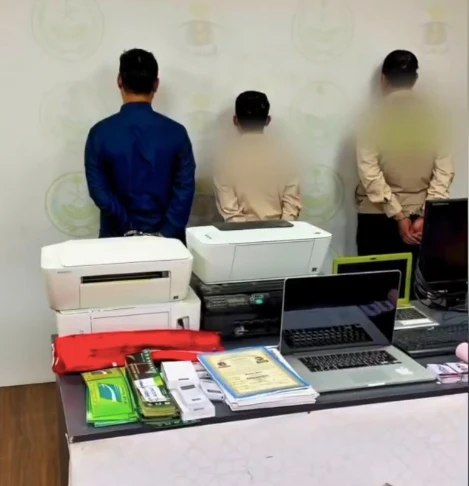 Three people with their backs to the camera stand behind a table with printers, a laptop, and office supplies in a room.
