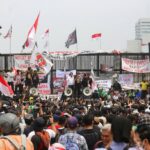 Large crowd of protesters gathered in front of a gated structure, waving banners and Indonesian flags with speakers on stage using megaphones