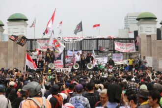 Large crowd of protesters gathered in front of a gated structure, waving banners and Indonesian flags with speakers on stage using megaphones