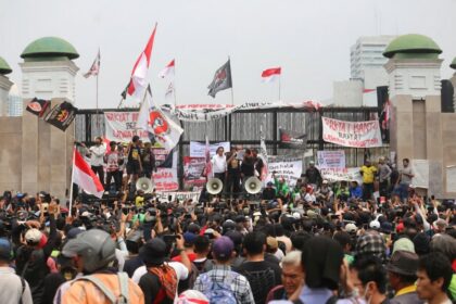 Large crowd of protesters gathered in front of a gated structure, waving banners and Indonesian flags with speakers on stage using megaphones