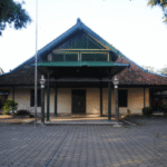 Traditional wooden building with a green, multi-tiered gabled roof and a covered front porch, set in a paved courtyard with trees framing the scene.