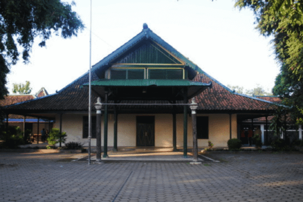 Traditional wooden building with a green, multi-tiered gabled roof and a covered front porch, set in a paved courtyard with trees framing the scene.