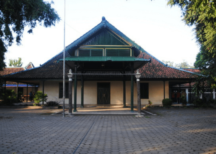 Traditional wooden building with a green, multi-tiered gabled roof and a covered front porch, set in a paved courtyard with trees framing the scene.