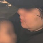 Close-up of a person in a dark hat kissing or nuzzling a baby whose face is blurred for privacy; soft lighting and dark clothing.