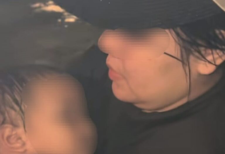 Close-up of a person in a dark hat kissing or nuzzling a baby whose face is blurred for privacy; soft lighting and dark clothing.